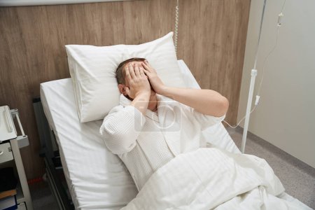 Téléchargez les photos : Portrait taille haute d'homme caucasien est couché sur le lit de la médecine avec ses mains sur les yeux dans la salle de médecine - en image libre de droit