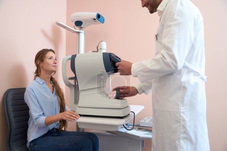 Photo for Side view portrait of beautiful Caucasian female is sitting on the chair while doctor is preparing eyesight machine in the clinic - Royalty Free Image