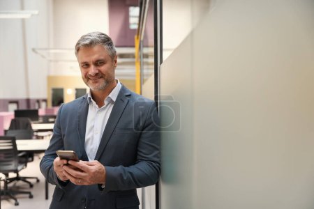 Photo for Adult man in office clothes standing in coworking near the wall, looking at mobile phone - Royalty Free Image