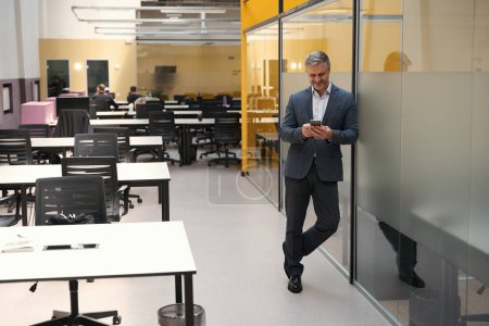 Foto de Hombre adulto en ropa de oficina de pie en coworking, mirando el teléfono móvil y charlando - Imagen libre de derechos