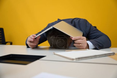 Téléchargez les photos : Homme adulte couché à la table se couvrant d'un ordinateur portable dans le bureau - en image libre de droit