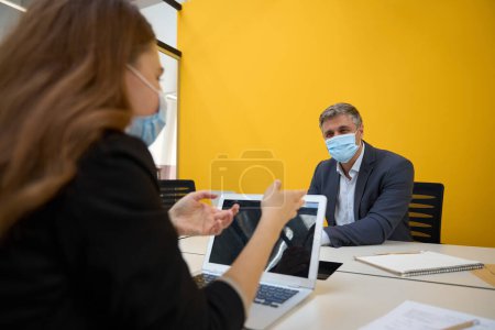 Téléchargez les photos : Femme et homme en vêtements de bureau et masques de protection assis à la table avec ordinateur portable et parler - en image libre de droit