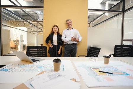 Foto de Hombre y mujer de pie junto a la mesa con planos arquitectónicos, hombre sosteniendo documento, mirando a la cámara - Imagen libre de derechos