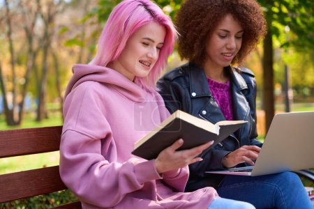 Téléchargez les photos : Souriant jeune femme lecture roman tandis que son compagnon féminin joyeux à l'aide d'un ordinateur portable - en image libre de droit
