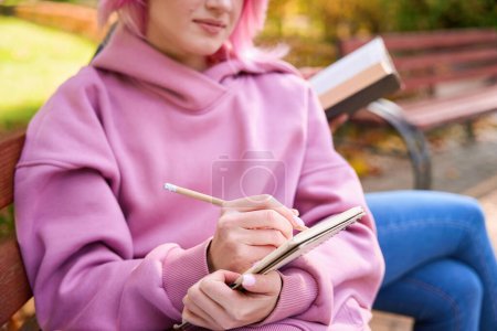 Téléchargez les photos : Photo recadrée d'une femme sérieuse écrivant au crayon dans un cahier ouvert assis sur un banc en bois à côté d'une personne avec un livre - en image libre de droit