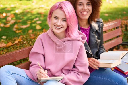 Téléchargez les photos : Souriant jeune femme écrit dans un cahier tout en étant assis sur le banc à côté de son ami heureux avec le manuel - en image libre de droit