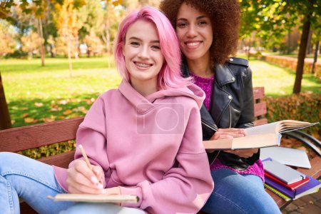 Photo for Smiling young female taking notes in notebook while sitting in bench next to her cheerful girlfriend with book - Royalty Free Image