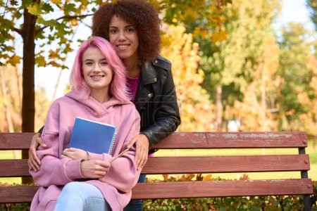 Photo for Smiling lady hugging her happy girlfriend seated on bench with textbooks in hands - Royalty Free Image
