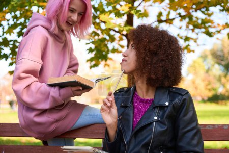 Téléchargez les photos : Dame joyeuse avec livre regardant son ami assis sur un banc en bois avec crayon à la main - en image libre de droit