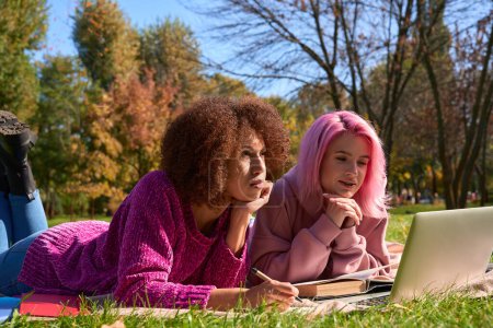 Téléchargez les photos : Jeune femme sérieuse avec stylo à la main couché sur l'herbe à côté de son compagnon féminin concentré avec livre - en image libre de droit