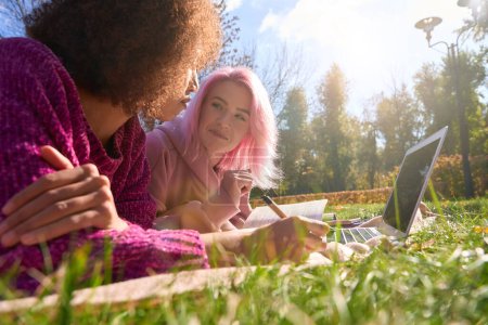 Téléchargez les photos : Femme avec crayon à la main couchée sur l'herbe à côté de son ami heureux avec le livre - en image libre de droit
