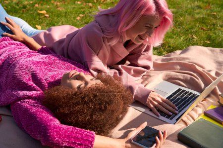 Foto de Señora tranquila con el teléfono celular acostado en la hierba junto a un amigo alegre utilizando el ordenador portátil - Imagen libre de derechos