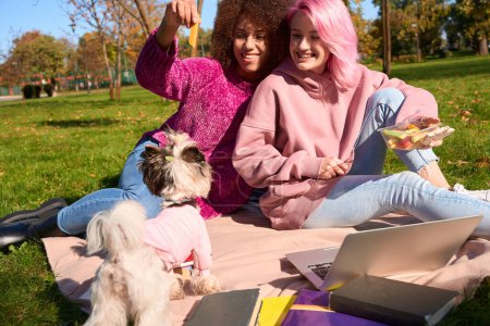 Foto de Mujer alegre con contenedor de comida en la mano sentada sobre una manta junto a su alegre amiga jugando con el perro - Imagen libre de derechos