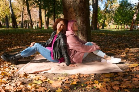 Téléchargez les photos : Souriante jeune femme et sa chère compagne assise sur une couverture sous un arbre - en image libre de droit