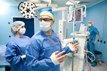 Téléchargez les photos : Processus de préparation à l'opération chirurgicale dans la salle préopératoire, l'infirmière aide le médecin à enfiler une robe stérile - en image libre de droit