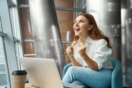 Téléchargez les photos : Joyeux jeune femme assise dans le bureau près de l'ordinateur portable, regardant vers le haut et se réjouit - en image libre de droit