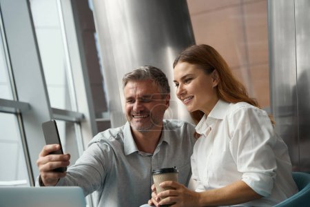 Foto de Feliz señora y hombre sentado en la oficina cerca de la computadora portátil, sosteniendo el teléfono móvil y tomando selfie - Imagen libre de derechos