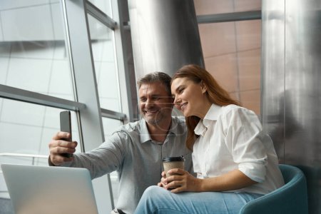 Foto de Mujer y hombre sentado en la oficina cerca de la computadora portátil, sosteniendo el teléfono móvil y tomando selfie - Imagen libre de derechos