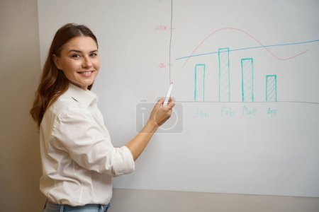 Photo for Young woman standing near board, holding marker and showing to camera schedule in the office - Royalty Free Image