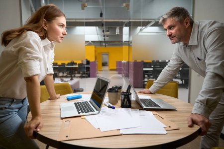 Photo for Young lady and man in office clothes standing near the table with laptop and looking at each other, conflict - Royalty Free Image
