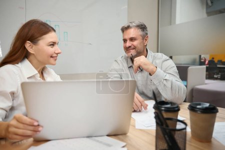 Téléchargez les photos : Dame assise à la table près d'un ordinateur portable, homme adulte en vêtements de bureau assis près et parlant - en image libre de droit