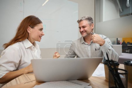 Téléchargez les photos : Architecte souriant assis à la table près de l'ordinateur portable, homme adulte en vêtements de bureau assis près et parlant - en image libre de droit