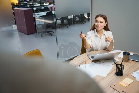 Foto de Mujer joven sentada junto a la mesa cerca del portátil en coworking y hablando con el empleado - Imagen libre de derechos