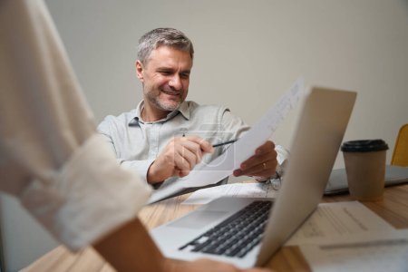 Téléchargez les photos : Homme adulte assis près de la table près de l'ordinateur portable en coworking, tenant le document et le donnant à l'employé - en image libre de droit