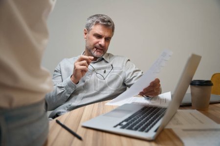 Téléchargez les photos : Homme adulte assis près de la table près d'un ordinateur portable en coworking, en tenant un document et en lisant - en image libre de droit