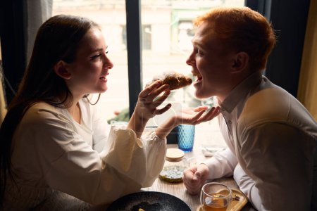 Téléchargez les photos : Jeune femme aux cheveux longs traite son mari avec un croissant, le couple est situé dans un café confortable près de la fenêtre - en image libre de droit