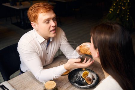Photo for Caring red-haired guy treats his girlfriend with a croissant, the couple is located in a cozy cafe by the window - Royalty Free Image
