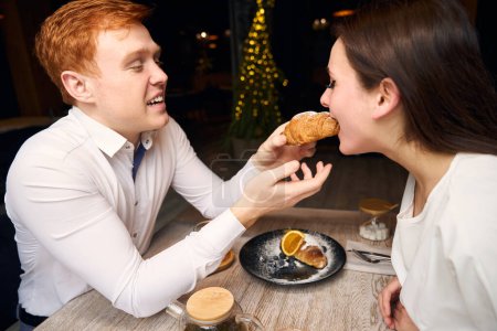 Téléchargez les photos : Beau roux traite sa petite amie avec un croissant, le couple est situé dans un café confortable près de la fenêtre - en image libre de droit