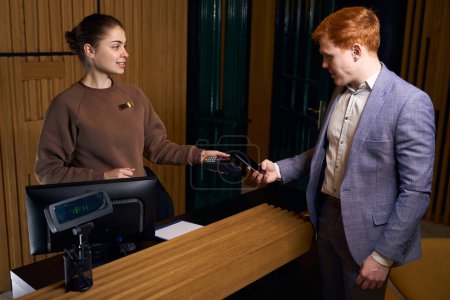 Photo for Young man pays for hotel services by phone, the administrator makes payment through the terminal - Royalty Free Image