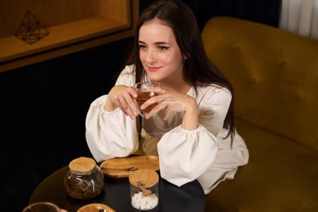 Téléchargez les photos : Jolie jeune brune boit du thé dans un salon d'hôtel, la femme est assise sur un canapé confortable - en image libre de droit