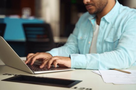 Téléchargez les photos : Jeune homme assis à table et tapant sur ordinateur portable pour le travail dans le centre d'affaires - en image libre de droit