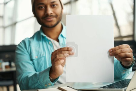 Téléchargez les photos : Homme en coiffure assis à l'ordinateur portable avec un morceau de papier dans ses mains, en regardant la caméra - en image libre de droit