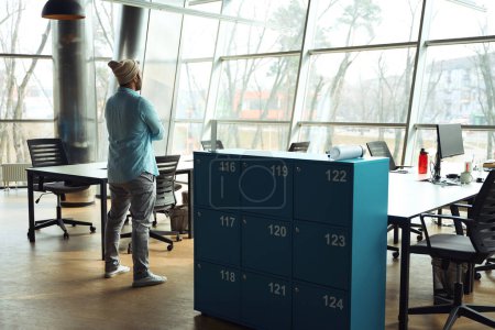 Téléchargez les photos : Jeune homme en chemise bleue debout dans le bureau près du placard, prêt pour le travail - en image libre de droit