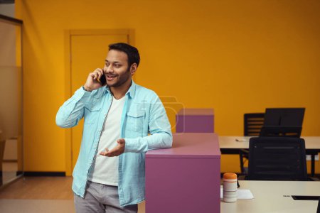 Foto de Sonriente hombre de pie cerca de la mesa y llamando por teléfono en el trabajo en el centro de negocios - Imagen libre de derechos
