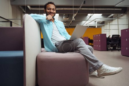 Téléchargez les photos : Homme souriant reposant et travaillant sur ordinateur dans le bureau, regardant la caméra - en image libre de droit