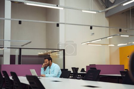 Téléchargez les photos : Jeune homme assis sur la chaise à la table et le travail sur ordinateur dans le centre d'affaires - en image libre de droit