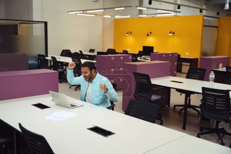 Téléchargez les photos : Heureux mâle assis sur la chaise au bureau devant l'ordinateur et lève les mains vers le haut dans le bureau - en image libre de droit