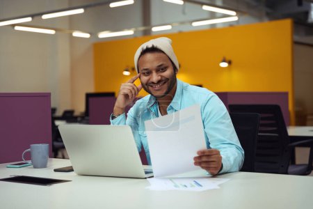 Téléchargez les photos : Heureux homme en chapeau assis au bureau devant un ordinateur portable et en regardant le document - en image libre de droit