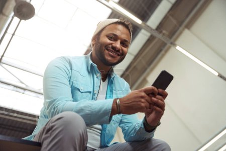 Téléchargez les photos : Homme souriant en chapeau assis dans le bureau et utilisant le téléphone, regardant la caméra - en image libre de droit