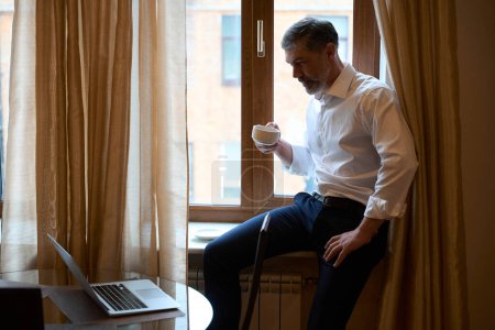 Téléchargez les photos : Homme assis sur le rebord de la fenêtre avec une tasse dans la main et regardant l'ordinateur portable - en image libre de droit