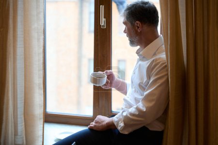 Téléchargez les photos : Homme en costume d'affaires avec une tasse à la main regardant par la fenêtre de l'hôtel - en image libre de droit