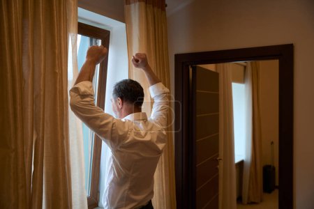 Téléchargez les photos : Homme adulte en vêtements de bureau debout près du rebord de la fenêtre et s'étirant dans le motel - en image libre de droit
