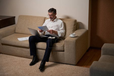 Téléchargez les photos : Homme adulte en tenue de travail assis sur le canapé, tenant un ordinateur portable et lisant des documents et travaillant - en image libre de droit