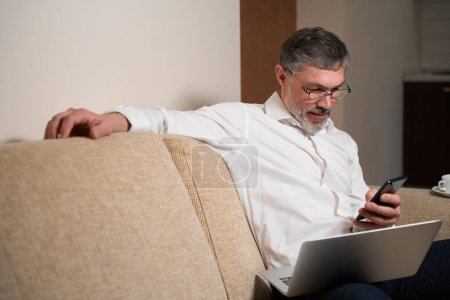 Téléchargez les photos : Homme adulte en tenue de travail assis sur le canapé, tenant un ordinateur portable, bavardant avec le client au téléphone, travaillant - en image libre de droit