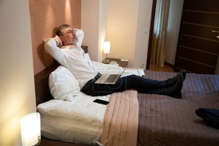 Téléchargez les photos : Homme professionnel en vêtements d'affaires assis sur le lit, tenant l'ordinateur portable et se reposant - en image libre de droit