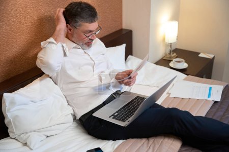 Téléchargez les photos : Travailleur professionnel en vêtements de travail assis sur le lit, tenant un ordinateur portable et lisant des documents - en image libre de droit